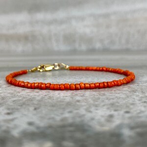 May include: A delicate orange beaded bracelet with a gold clasp. The bracelet is made of small, round beads and is perfect for layering with other bracelets.