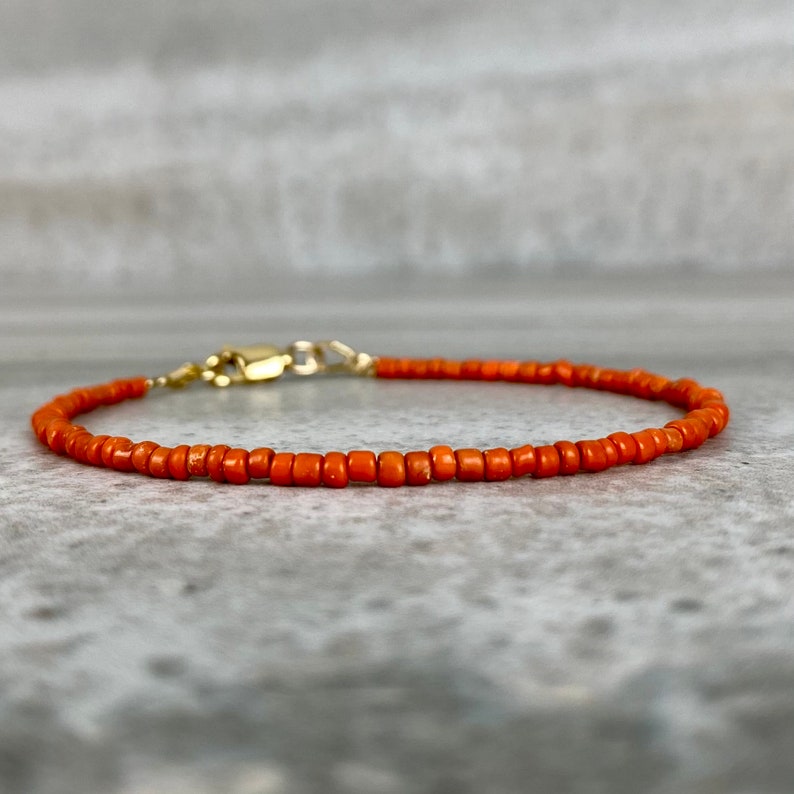 May include: A delicate orange beaded bracelet with a gold clasp. The bracelet is made of small, round beads and is perfect for layering with other bracelets.