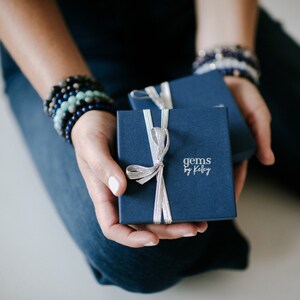 May include: A square navy blue gift box with a silver ribbon tied in a bow. The text "gems by Kelley" is printed in silver on the lid. The box is held in place with a second matching navy blue box visible behind it, featuring a silver ribbon.