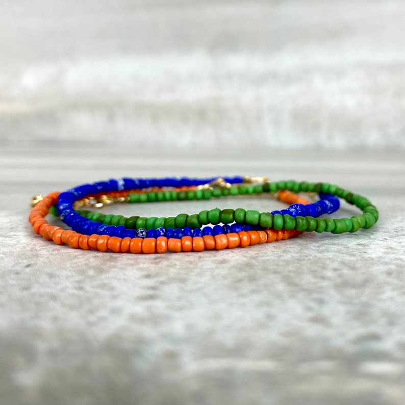 May include: A layered bracelet made of small, colourful beads. The bracelet is made of three strands of beads: orange, green, and blue.