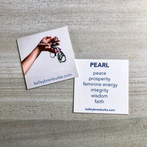 May include: A white card with the word "PEARL" in blue text. Below the word "PEARL" are the words "peace", "prosperity", "feminine energy", "integrity", "wisdom", and "faith" in blue text. The card also includes the website "kelleybrenburke.com" in blue text. A second card with a hand holding a strand of pearls is visible in the background.
