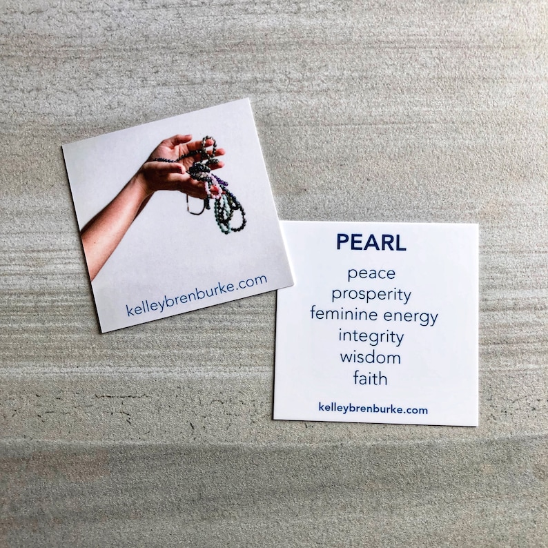 May include: A white card with the word "PEARL" in blue text. Below the word "PEARL" are the words "peace", "prosperity", "feminine energy", "integrity", "wisdom", and "faith" in blue text. The card also includes the website "kelleybrenburke.com" in blue text. A second card with a hand holding a strand of pearls is visible in the background.