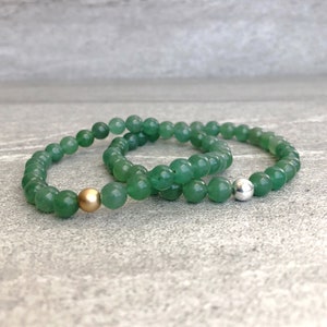 May include: Two beaded bracelets made of smooth, round green aventurine stones. One bracelet features a single matte gold-toned metal bead, while the other bracelet includes a single polished silver-toned metal bead as a focal point.