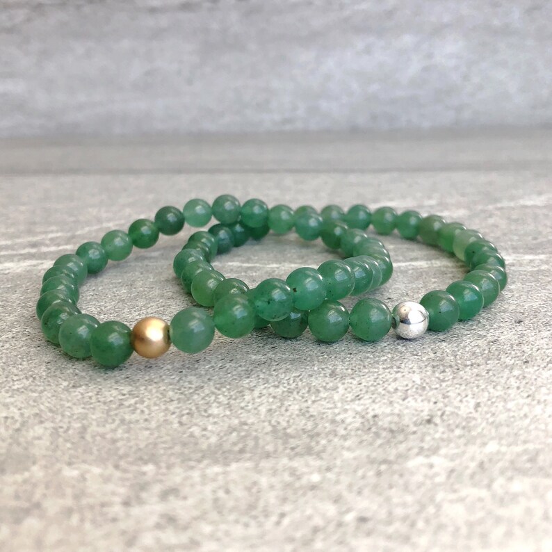 May include: Two beaded bracelets made of smooth, round green aventurine stones. One bracelet features a single matte gold-toned metal bead, while the other bracelet includes a single polished silver-toned metal bead as a focal point.