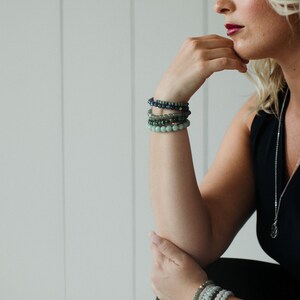 May include: A stack of beaded bracelets worn on each wrist. The upper stack features dark blue, forest green, and light green spherical beads. The lower stack consists of light green, grey, and white spherical beads, each featuring a small metallic accent.