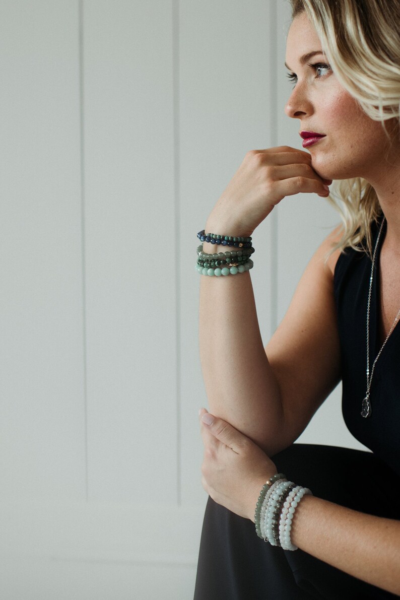 May include: A stack of beaded bracelets worn on each wrist. The upper stack features dark blue, forest green, and light green spherical beads. The lower stack consists of light green, grey, and white spherical beads, each featuring a small metallic accent.