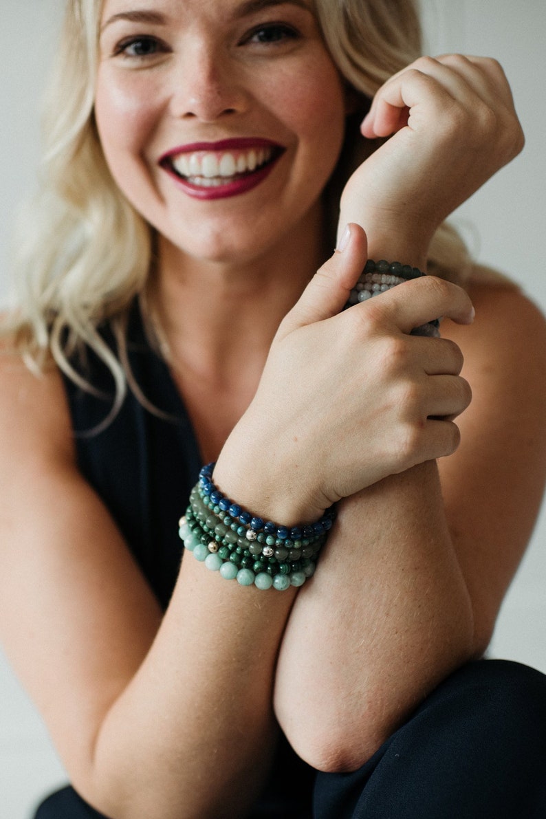 May include: A stack of five beaded bracelets featuring round gemstone beads in varying shades of blue and green, including dark blue, forest green, and light mint green, with small metallic accent beads interspersed throughout the design.