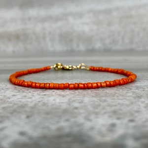 May include: A delicate orange beaded bracelet with a gold clasp. The bracelet is made of small, round beads and is perfect for layering with other bracelets.