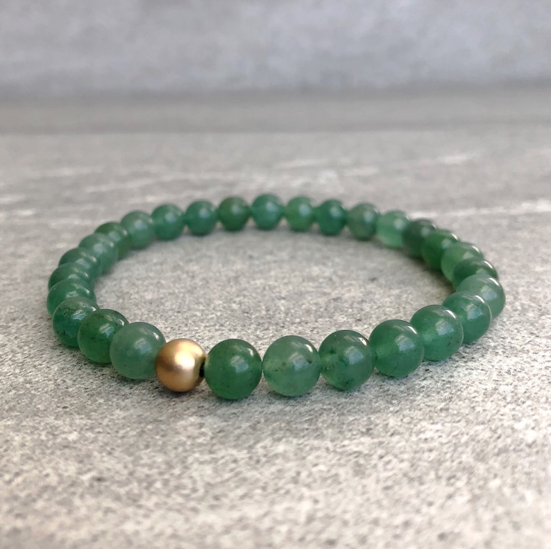 May include: A stretch bracelet composed of smooth, round green aventurine beads. The circular arrangement features a single, polished gold-tone metal bead positioned as a focal point among the uniform green gemstone beads.