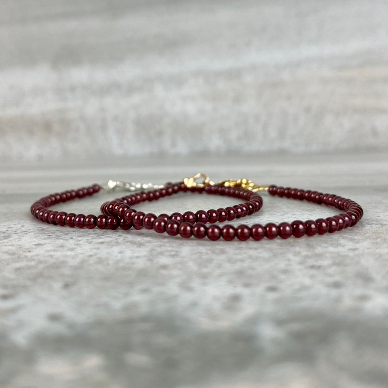 May include: Two maroon beaded bracelets with gold and silver clasps. The bracelets are on a gray textured surface.