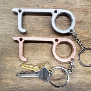 May include: Two door handle openers, one silver and one rose gold, attached to a silver key chain with a key ring. The key chain has a small silver tag with the text "VERMONT CRAFT CO."