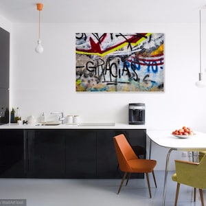 K&ouml;nnte beinhalten: Eine moderne K&uuml;che mit schwarzen Schr&auml;nken und einer wei&szlig;en Arbeitsplatte. Ein farbenfroher Graffiti-Kunstdruck h&auml;ngt an der Wand &uuml;ber einem wei&szlig;en Esstisch mit vier St&uuml;hlen. Der Tisch ist mit einer Sch&uuml;ssel roten Tomaten gedeckt.