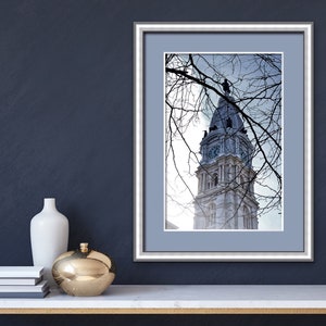 K&ouml;nnte beinhalten: Ein gerahmtes Foto eines hohen, wei&szlig;en Geb&auml;udes mit einem Uhrturm. Das Geb&auml;ude ist teilweise von kahlen &Auml;sten verdeckt. Das Foto ist in einem silbernen Rahmen mit einem hellblauen Passepartout gerahmt.