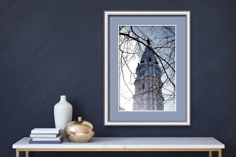 K&ouml;nnte beinhalten: Ein gerahmtes Foto eines hohen, wei&szlig;en Geb&auml;udes mit einem Uhrturm. Das Geb&auml;ude ist teilweise von kahlen &Auml;sten verdeckt. Das Foto ist in einem silbernen Rahmen mit einem hellblauen Passepartout gerahmt.