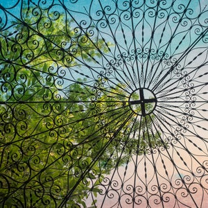 May include: A close-up view of a black metal gazebo roof with intricate scrollwork designs. The roof is made of a series of curved metal bars that create a pattern of circles and swirls. The roof is open to the sky, and the sky is a light blue colour with some clouds. The roof is surrounded by green trees.