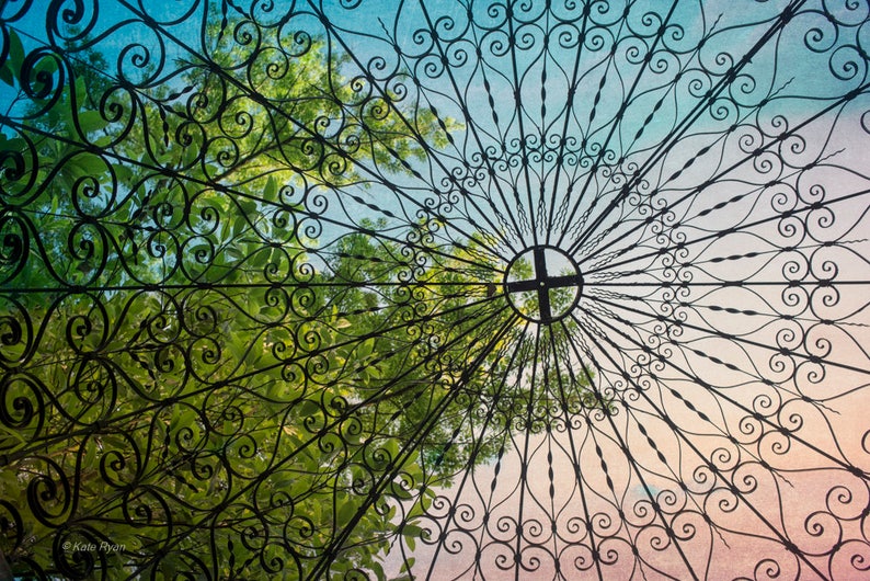 May include: A close-up view of a black metal gazebo roof with intricate scrollwork designs. The roof is made of a series of curved metal bars that create a pattern of circles and swirls. The roof is open to the sky, and the sky is a light blue colour with some clouds. The roof is surrounded by green trees.