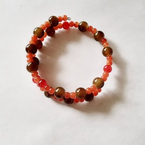 May include: A spiral bracelet featuring alternating rows of brown and orange beads. The bracelet is made of small, round beads, with larger, darker brown beads interspersed throughout. The color palette includes shades of brown, orange, and red.