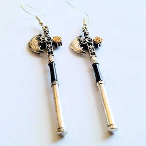 May include: A pair of silver-toned axe-shaped earrings with black and gold accents. The earrings feature a silver axe head, a black central section, and a silver handle. A small gold-colored rose is at the top of each earring.