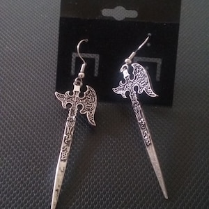 May include: A pair of silver-colored earrings shaped like stylized axes. The earrings have intricate detailing on the axe heads and handles, with a hook for the ear. They are displayed on a black card.