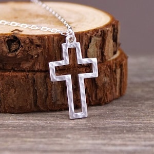 May include: A silver-colored cross pendant necklace. The cross has a hammered texture and is suspended from a delicate chain. The necklace is displayed on a wooden surface, showcasing its design and craftsmanship.