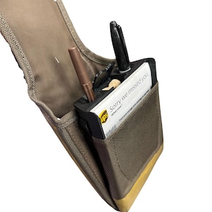 Puede incluir: Una cartera de lona marrón y beige con un bolígrafo negro, un bolígrafo marrón y un albarán de entrega de UPS que dice "Sorry we missed you". La cartera tiene un acento dorado en la parte inferior.