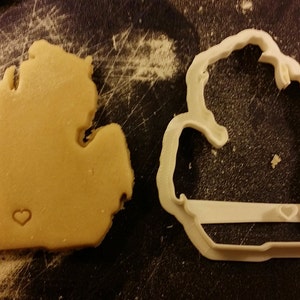 May include: A cookie in the shape of the state of Michigan with a small heart cutout. Next to it is a white plastic cookie cutter in the shape of Michigan with a heart cutout.
