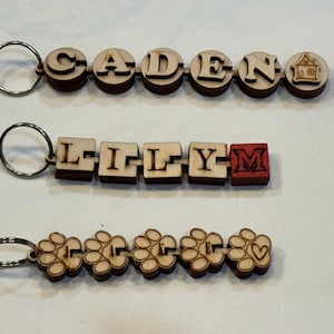 May include: Three wooden keychains. The top keychain spells "CADEN" with a house icon. The middle keychain spells "LILLY" with a red "M". The bottom keychain features flower-shaped charms.