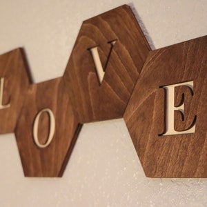 May include: Wooden wall decor spelling out the word "LOVE" in white letters on a dark brown background. Each letter is on a separate hexagonal piece of wood.