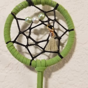 May include: A green dreamcatcher with black string and white and green beads. The dreamcatcher has a tassel hanging from the bottom.