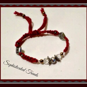 Puede incluir: Pulsera de cuerda roja con cuentas plateadas y doradas y detalles de cristal transparente. La pulsera tiene un cierre de nudo deslizante. "Sophisticated Trends" está escrito en el fondo blanco.