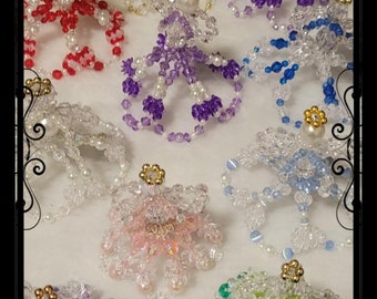 Op de afbeelding: Assortiment van kralenengel-versieringen in verschillende kleuren, waaronder rood, paars, blauw, groen en helder. Elke versiering heeft een kralenhalo en een kralenjurk.