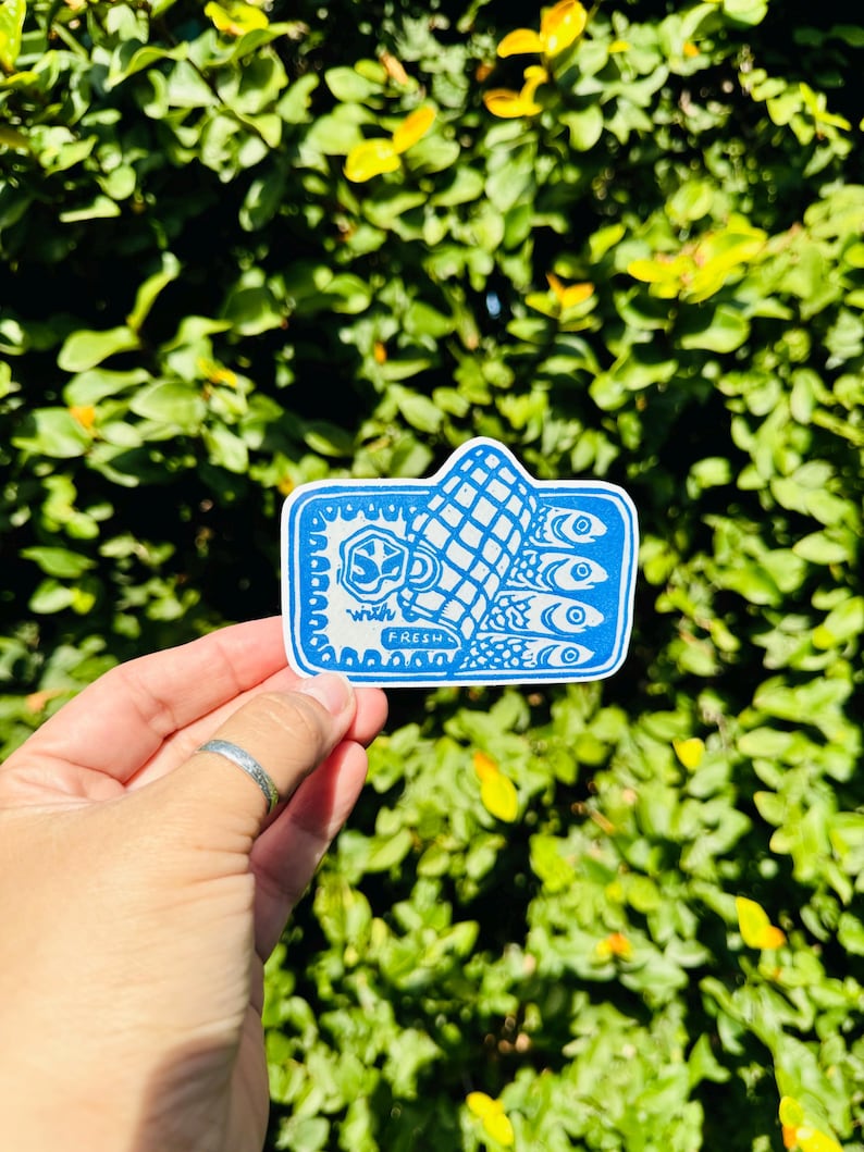 Op de afbeelding: Een blauwe en witte sticker met een hand die een vis vasthoudt. De hand houdt een vis vast met de tekst "with fresh" erop.