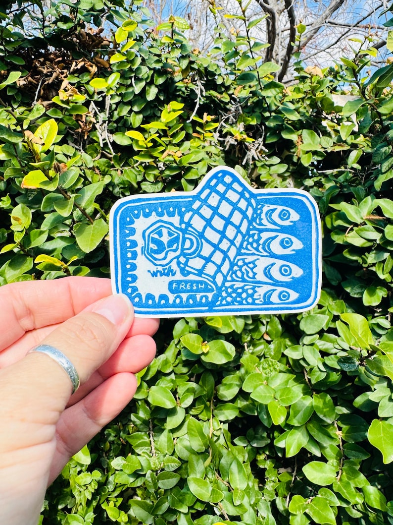 Op de afbeelding: Een blauwe en witte sticker met een ontwerp van een blikje sardines en de tekst "with fresh".