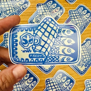 Op de afbeelding: Blauw-witte sticker met een hand die drie vissen vasthoudt en de tekst "with fresh" op een houten oppervlak.