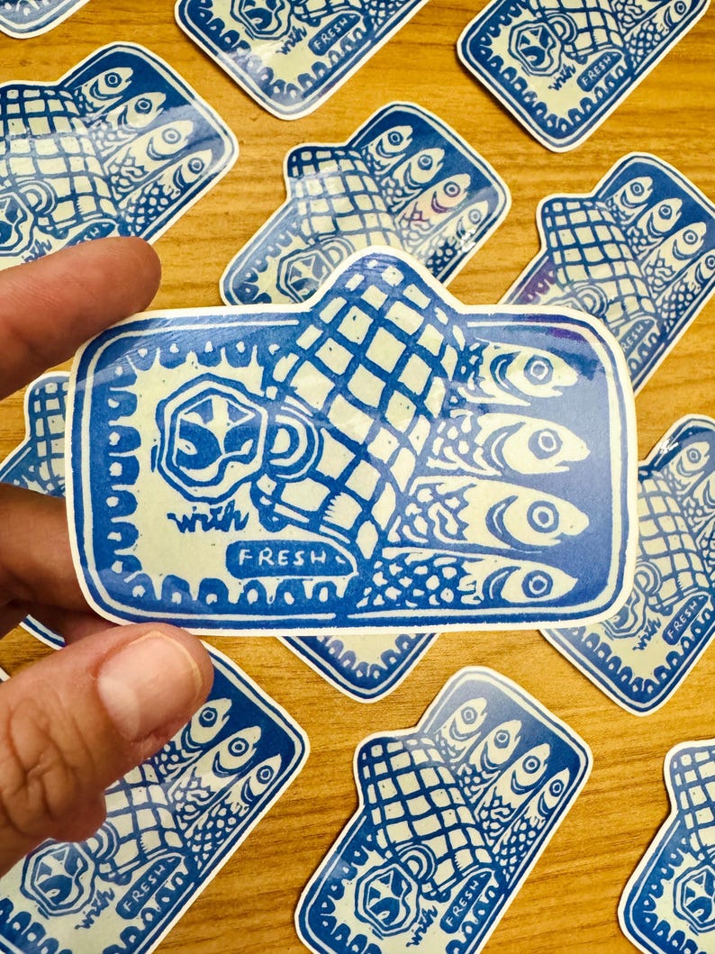 Op de afbeelding: Blauw-witte sticker met een hand die drie vissen vasthoudt en de tekst "with fresh" op een houten oppervlak.