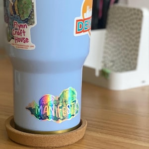 Op de afbeelding: Een lichtblauwe tumbler met een kurken onderzetter. De tumbler heeft verschillende stickers, waaronder een met de tekst "Flynn Craft House" en een andere met de tekst "DENVER". Er is ook een holografische sticker met de tekst "MANIFEST!"