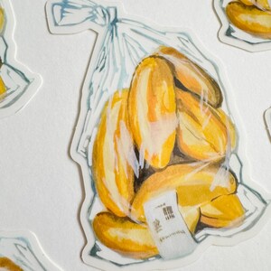 Op de afbeelding: Een aquarel-illustratie van een plastic zak gevuld met gele en oranje gelukkoekjes. De zak is bovenaan dichtgeknoopt en heeft een wit label met zwarte tekst.