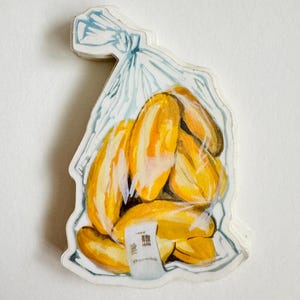 Op de afbeelding: Een aquarel-illustratie van een plastic zak gevuld met gele bananen. De zak is bovenaan dichtgeknoopt en heeft een klein wit label met zwarte tekst.