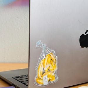 Op de afbeelding: Een doorzichtige plastic zak met een tros gele bananen erin. De zak zit op een zilveren laptop.