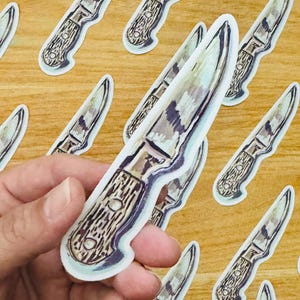May include: A close-up of a hand holding a sticker of a vintage-style knife with a wooden handle. The sticker is white with a light blue outline and is printed with a watercolor-style design. The sticker is on a wooden surface with other similar stickers.
