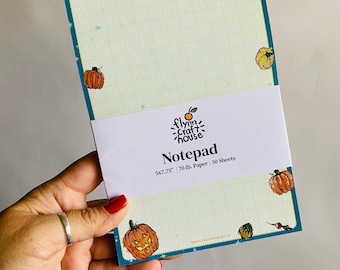 May include: A teal notepad with a Halloween theme. The notepad has a grid pattern and features illustrations of pumpkins with jack-o'-lantern faces. The notepad is 5x7.75 inches and has 70 lb. paper with 50 sheets. The text "flynn craft house" and "Notepad" are printed on the front of the notepad.