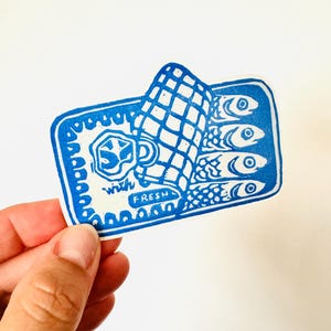 Op de afbeelding: Blauw-witte sticker van een blikje sardines met de tekst "with fresh" aan de onderkant. Het blikje is open en de sardines zijn zichtbaar.
