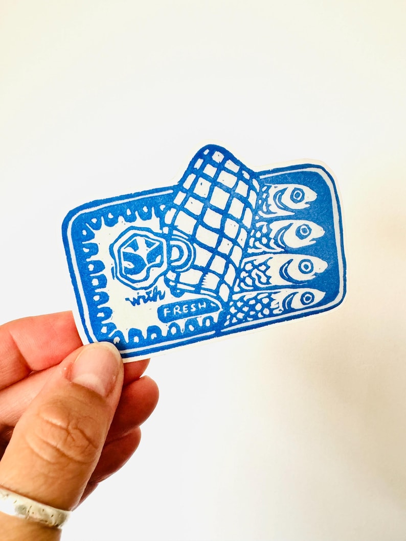 Op de afbeelding: Blauw-witte sticker van een blikje sardines met de tekst "with fresh" aan de onderkant. Het blikje is open en de sardines zijn zichtbaar.