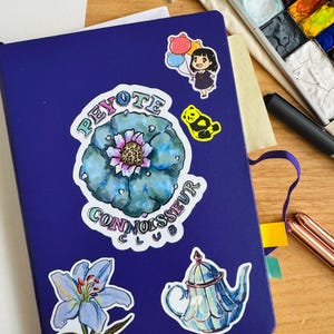 Op de afbeelding: Een blauw notitieboek met verschillende stickers, waaronder een cactus met de tekst "Peyote Connoisseur Club", een blauwe bloem, een theepot en een cartoonfiguur met ballonnen.