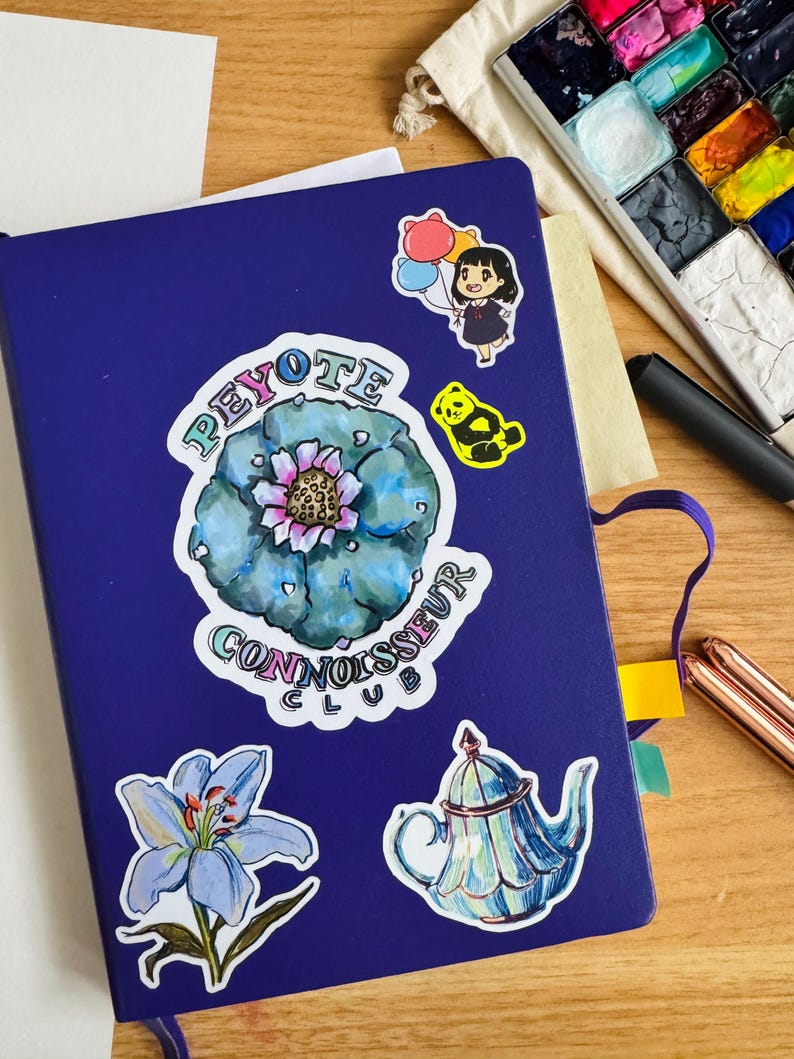 Op de afbeelding: Een blauw notitieboek met verschillende stickers, waaronder een cactus met de tekst "Peyote Connoisseur Club", een blauwe bloem, een theepot en een cartoonfiguur met ballonnen.