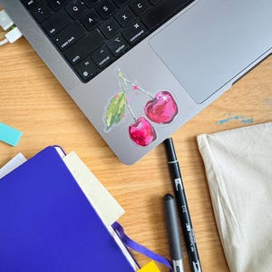Op de afbeelding: Een blauw notitieboek met een gele sticky note en een wit oppervlak met een laptop, twee pennen en een sticker met twee rode kersen met groene bladeren.