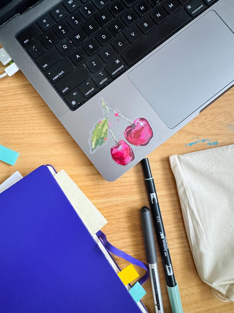 Op de afbeelding: Een blauw notitieboek met een gele sticky note en een wit oppervlak met een laptop, twee pennen en een sticker met twee rode kersen met groene bladeren.