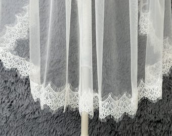 May include: A sheer white veil with delicate lace trim along the edges. The lace features an intricate floral design, adding a touch of elegance. The veil is draped, showcasing its lightweight and flowing fabric.