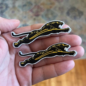 May include: Two embroidered patches featuring a black panther in a running pose. The panther has yellow stripes and details, with a white border. The patches are held in a hand, showcasing the design and craftsmanship.
