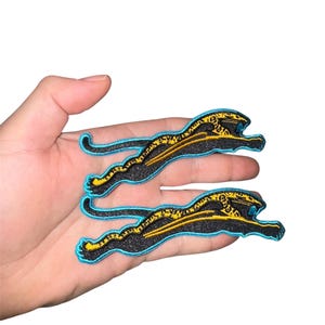 May include: Two black and gold cheetah patches with a turquoise outline. The patches are embroidered and have a detailed design.